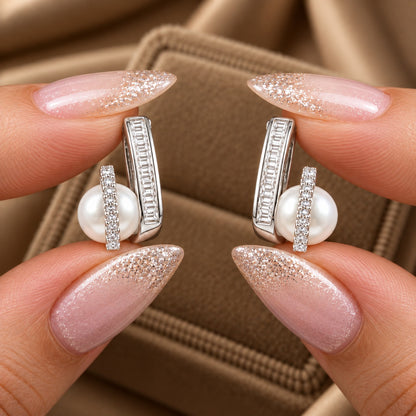 Pair of silver-tone pearl stud earrings held between manicured fingers, showing round zircon and baguette-cut stone detailing