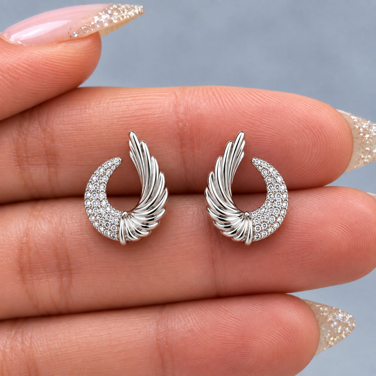 Pair of silver Celestial Wing Crescent Studs displayed on fingers, showing the size, clear zircon setting and detailed wing shape