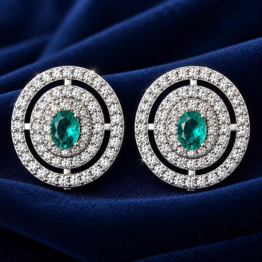 Close-up of round silver-tone earrings featuring an emerald-green oval centre stone surrounded by multiple rows of clear zircon.