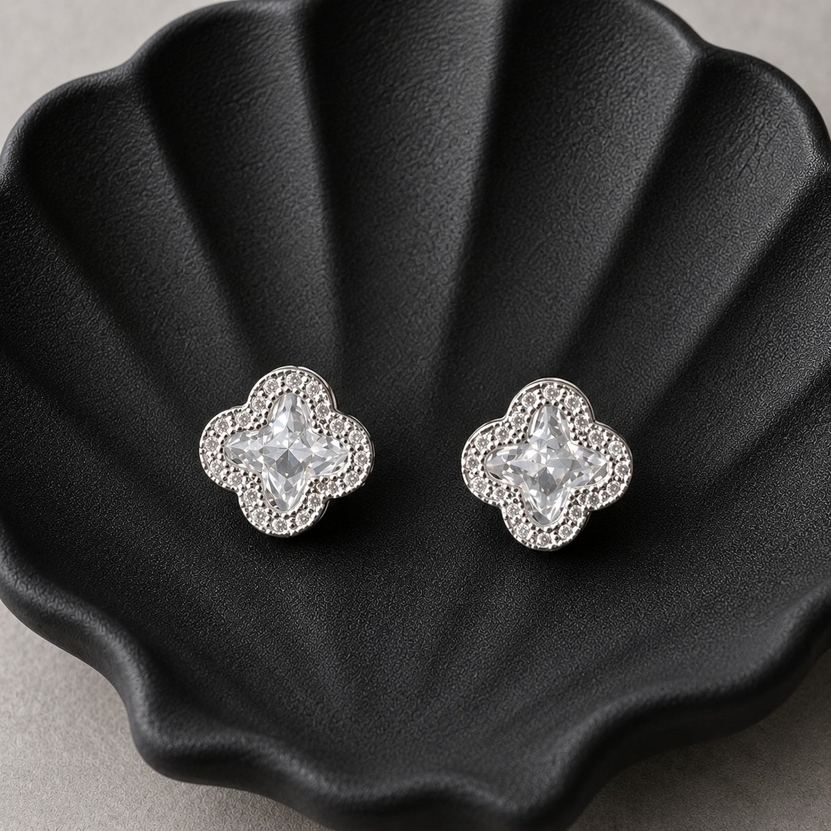 S925 sterling silver quatrefoil stud earrings with faceted clear centre crystals and zircon borders displayed on a black shell-shaped tray.