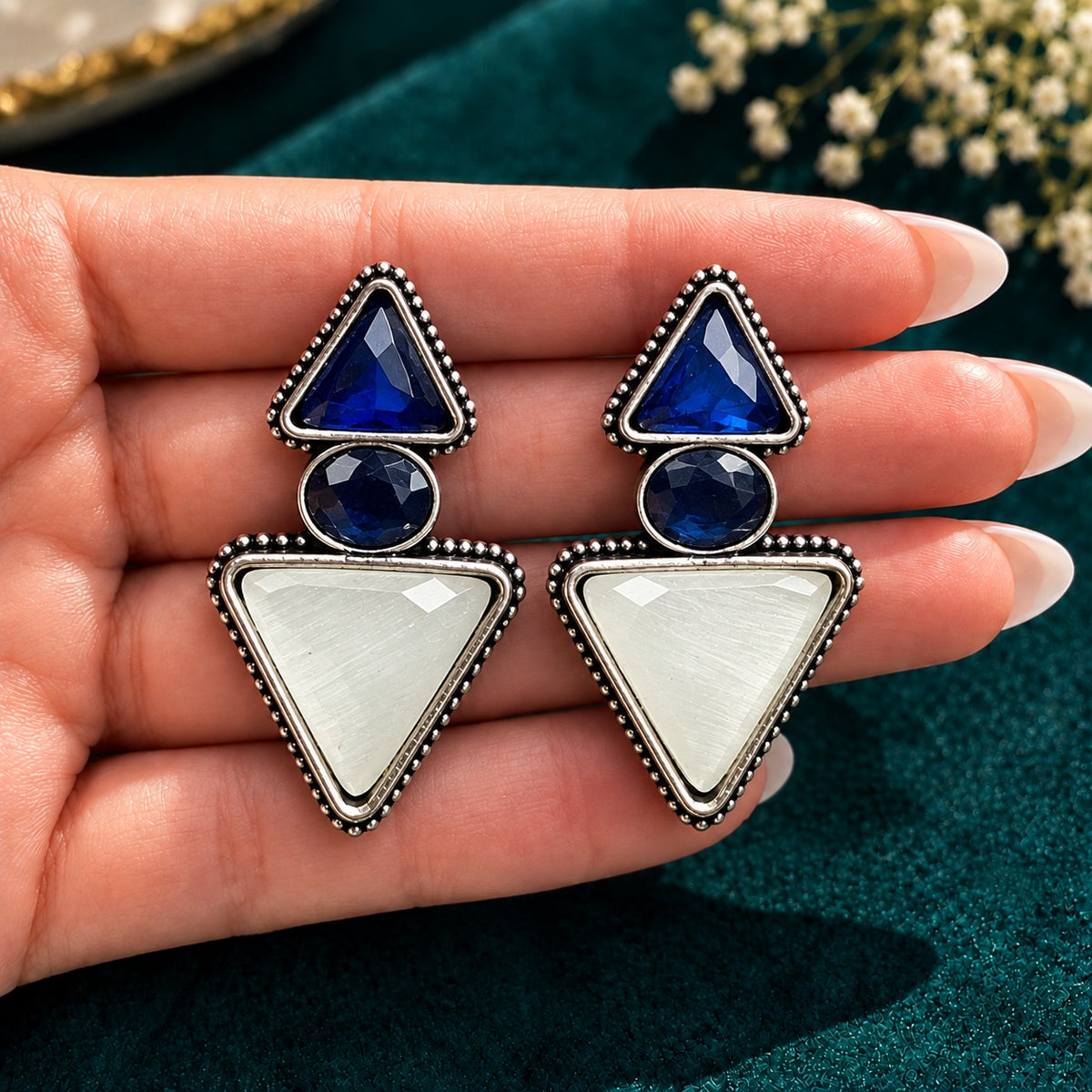 Sapphire blue and ivory Legacy Trine Earrings with opposing triangular stones, oval centre and silver-tone beaded setting on a hand