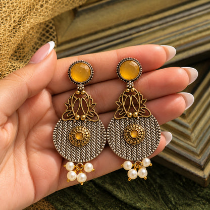 The Loom & Lotus Earrings displayed on a hand, showing their statement length, woven metal texture, mixed-metal detailing and pearl-drop finish.