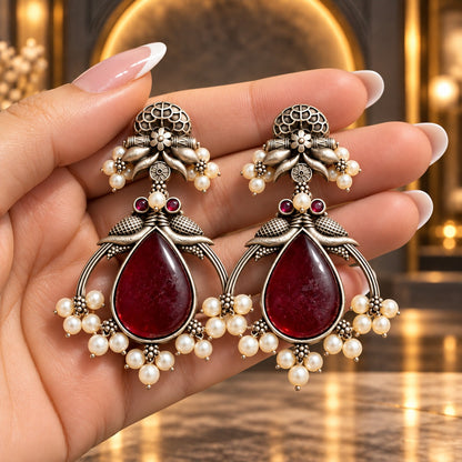 Hand-held close-up of The Royal Treasury Earrings showing their size, ornate oxidised silver-tone metalwork and pearl-like clusters.