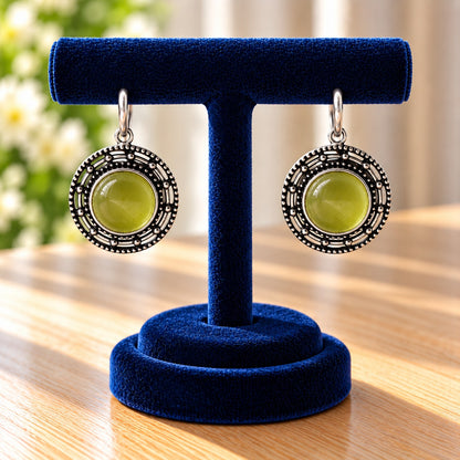 Circular medallion earrings featuring a luminous olive-lime-green centre surrounded by geometric openwork and antique silver-tone detailing.