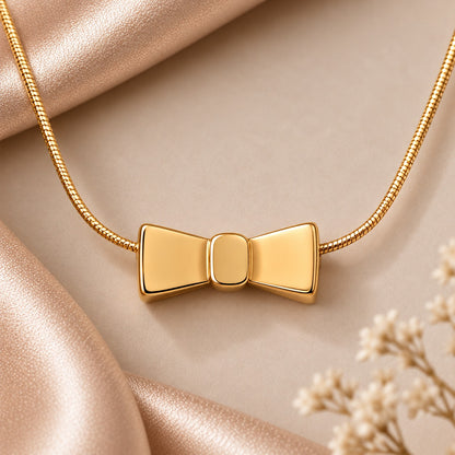 Close-up of a polished golden bow-shaped locket with smooth tapered wings and a rounded central knot, suspended from a delicate gold-tone box chain against a champagne satin background.