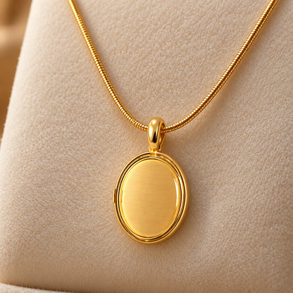 Gold-tone oval locket pendant with a smooth centre, suspended from a fine chain on a soft ivory jewellery stand.