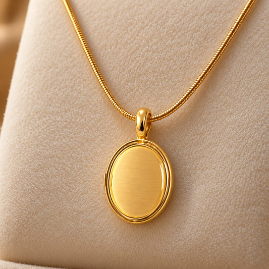 Gold-tone oval locket pendant with a smooth centre, suspended from a fine chain on a soft ivory jewellery stand.