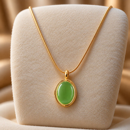 Gold-tone oval locket pendant with a smooth muted emerald green centre, suspended from a fine chain on a soft ivory jewellery stand.