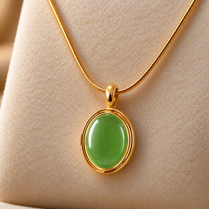 Gold-tone oval locket pendant with a smooth muted emerald green centre, suspended from a fine chain on a soft ivory jewellery stand.