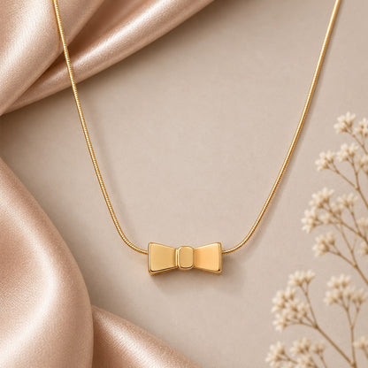 polished golden bow-shaped locket with smooth tapered wings and a rounded central knot, suspended from a delicate gold-tone box chain against a champagne satin background.