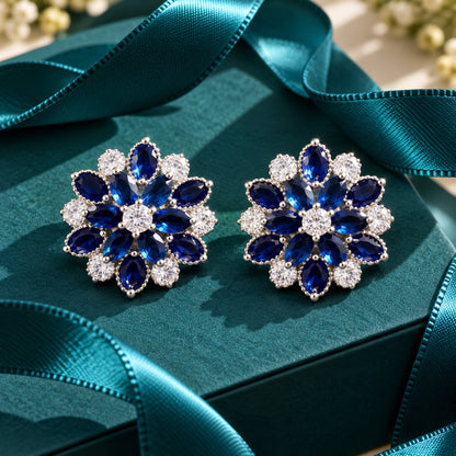 Vespera Bloom Earrings in sapphire blue, featuring layered marquise-cut gemstones, clear zircon accents and polished silver-tone settings, displayed on a teal jewellery box.