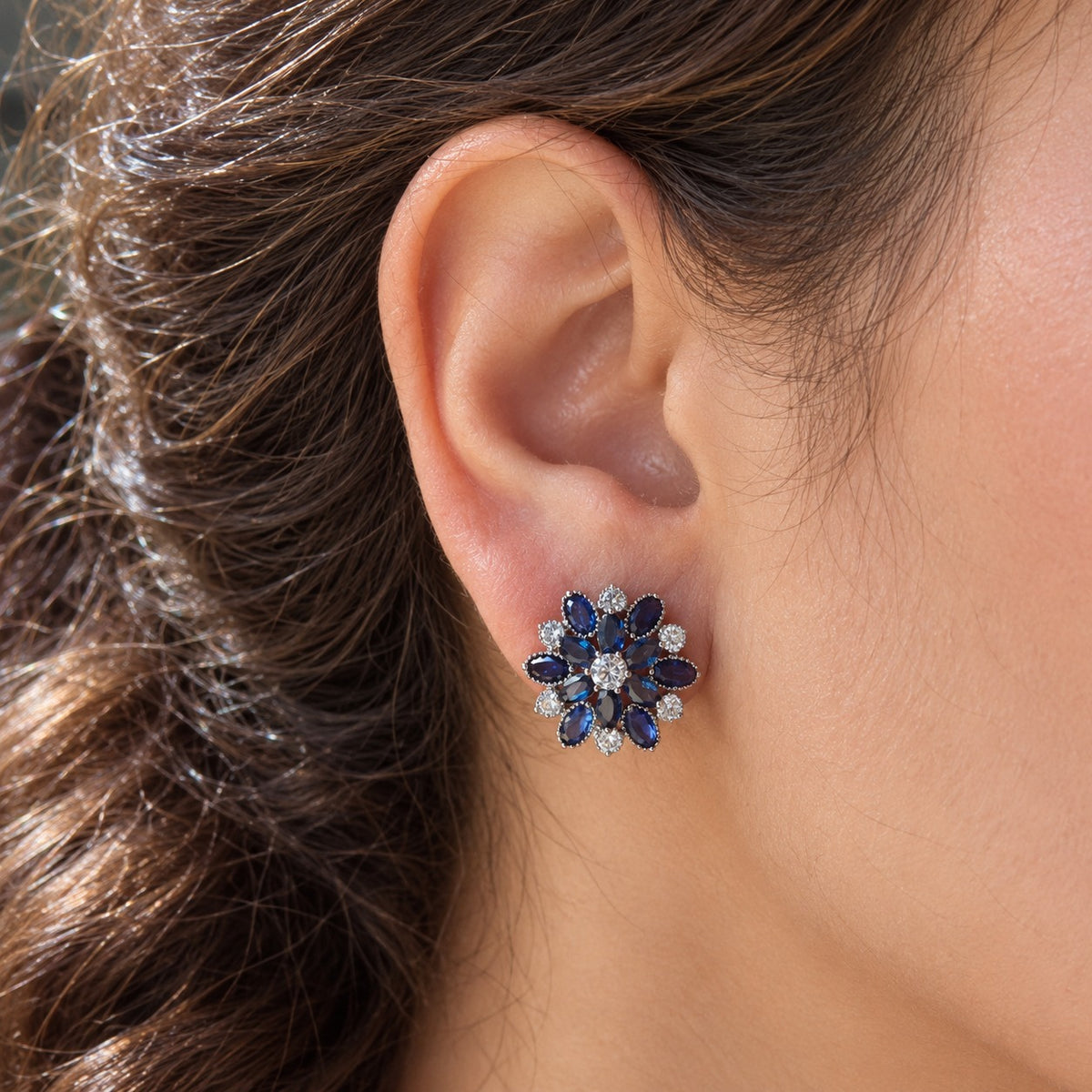 woman's ear wearing the Vespera Bloom sapphire blue floral stud earring, featuring vivid red gemstone petals and brilliant clear zircon detailing.