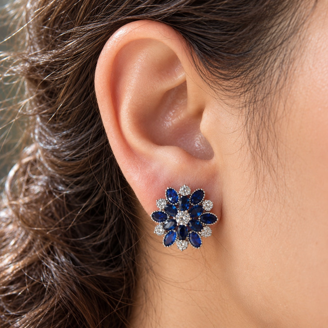 Close-up of a woman's ear wearing the Vespera Bloom sapphire blue floral stud earring, featuring vivid red gemstone petals and brilliant clear zircon detailing.