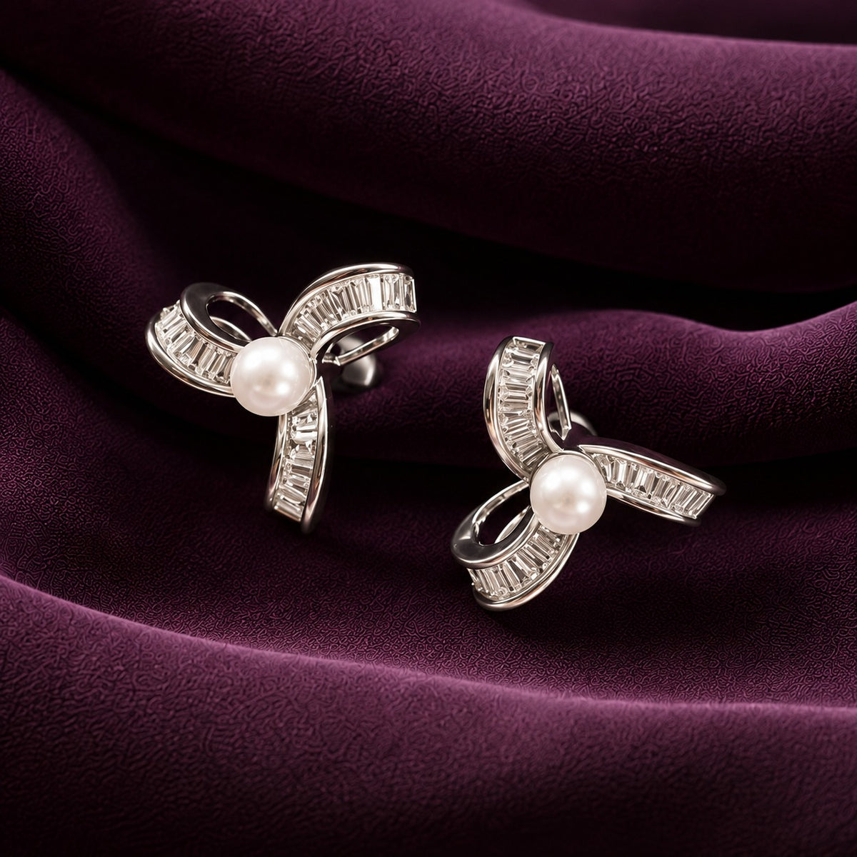 Silver-tone ribbon swirl stud earrings with white pearl centres and baguette-cut zircon stones, displayed on deep plum fabric.