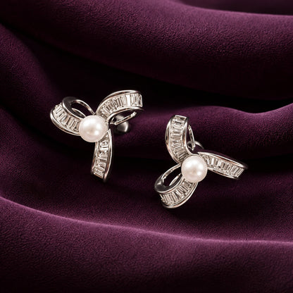 Silver-tone ribbon swirl stud earrings with white pearl centres and baguette-cut zircon stones, displayed on deep plum fabric.