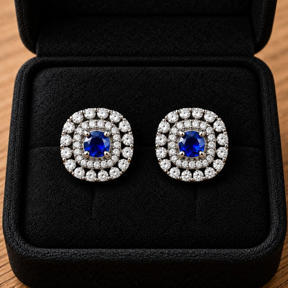 Close-up of silver-tone cushion-shaped stud earrings featuring deep sapphire blue faceted centre stones and intricate double-halo zircon detailing.
