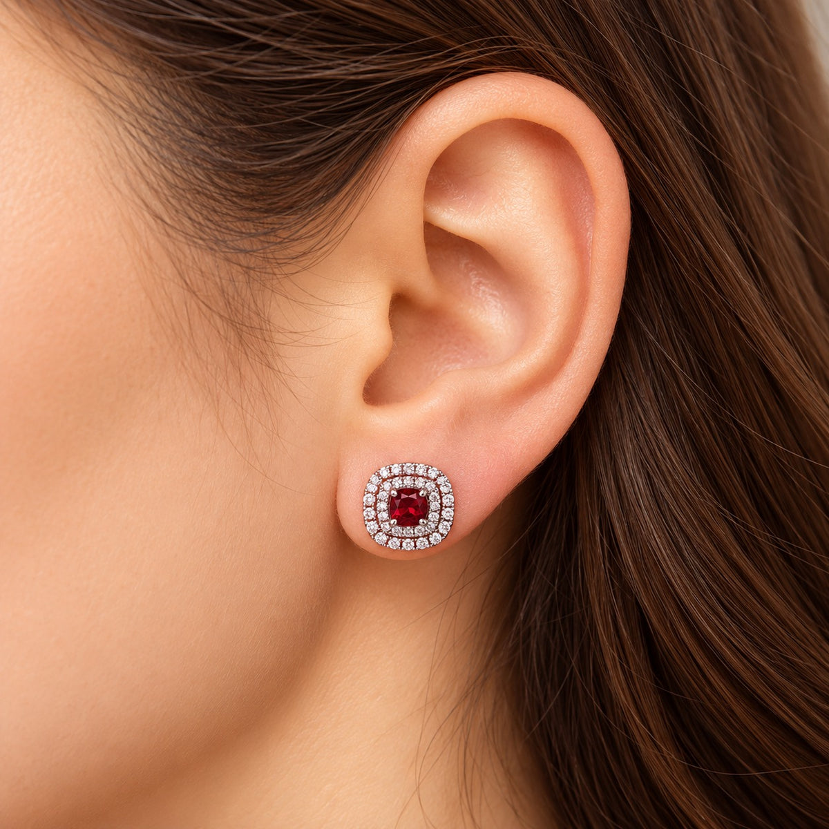 Close-up of a woman's ear wearing the Regent Cushion Zircon Stud in ruby red, highlighting its cushion-shaped design and clear zircon borders.