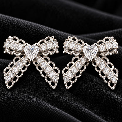 Close-up of Amorette Bow Studs featuring clear heart-shaped centre stones, clear zircon detailing, and polished silver-tone bow designs on black fabric.