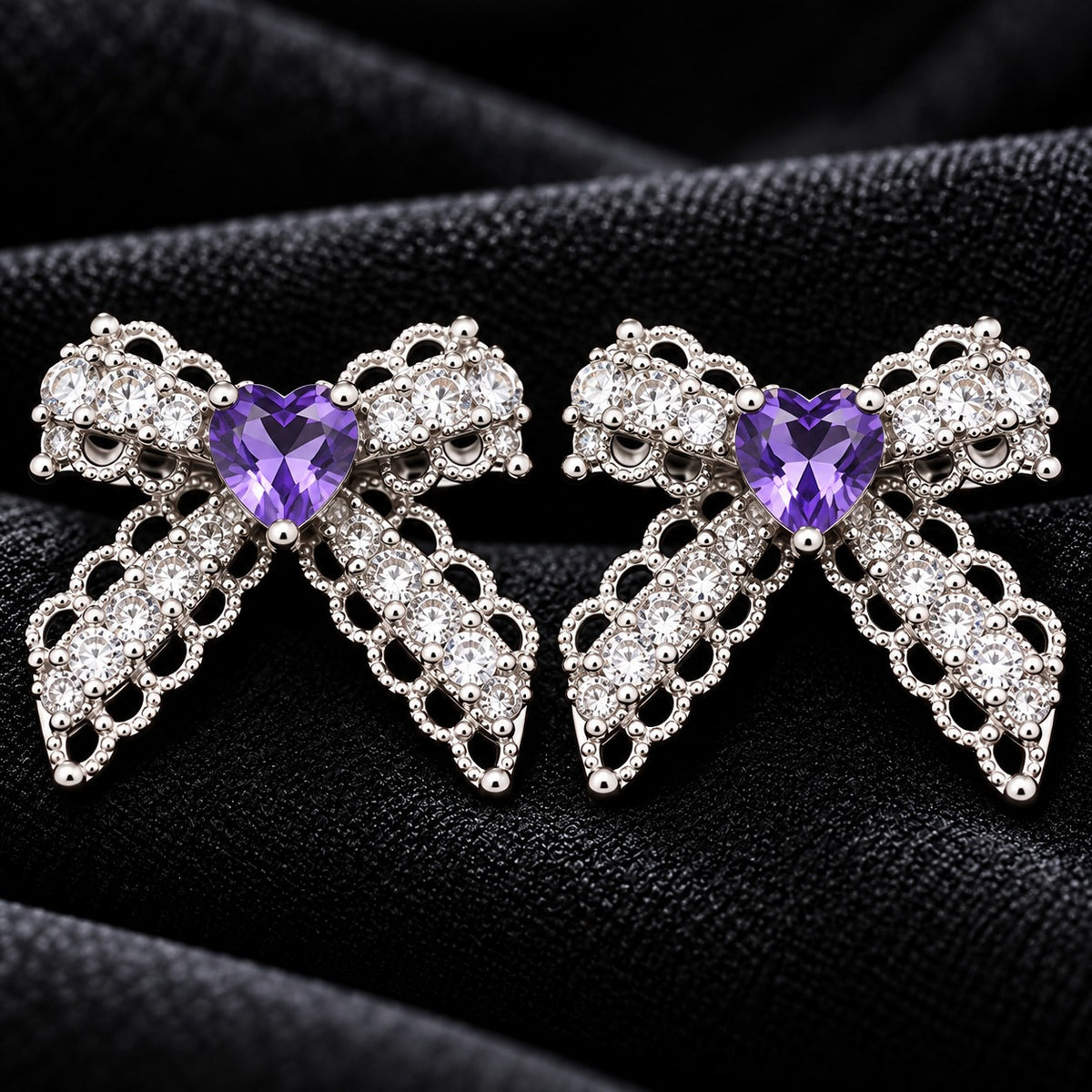 Close-up of Amorette Bow Studs featuring amethyst purple heart-shaped centre stones, clear zircon detailing, and polished silver-tone bow designs on black fabric.