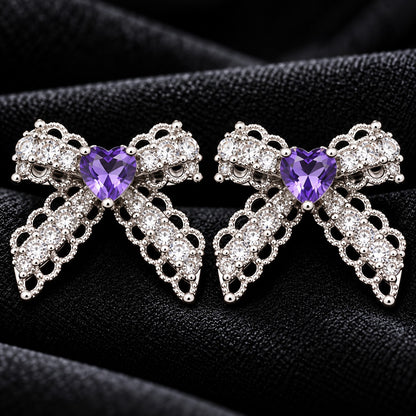 Close-up of Amorette Bow Studs featuring amethyst purple heart-shaped centre stones, clear zircon detailing, and polished silver-tone bow designs on black fabric.