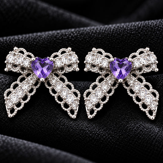 Close-up of Amorette Bow Studs featuring amethyst purple heart-shaped centre stones, clear zircon detailing, and polished silver-tone bow designs on black fabric.