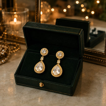 A pair of gold drop earrings with large clear teardrop gemstones and smaller round clear stones, displayed in an open black jewelry box on a marble surface with warm blurred lights in the background.