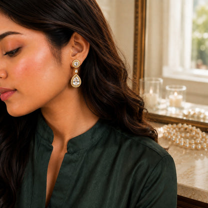 A woman wearing a dangling teardrop earring with a silver-toned round setting and clear teardrop gemstone, shown in profile against an indoor background with candles and pearls.