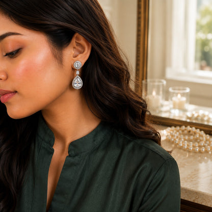 A woman wearing a dangling teardrop earring with a silver-toned round setting and clear teardrop gemstone, shown in profile against an indoor background with candles and pearls.