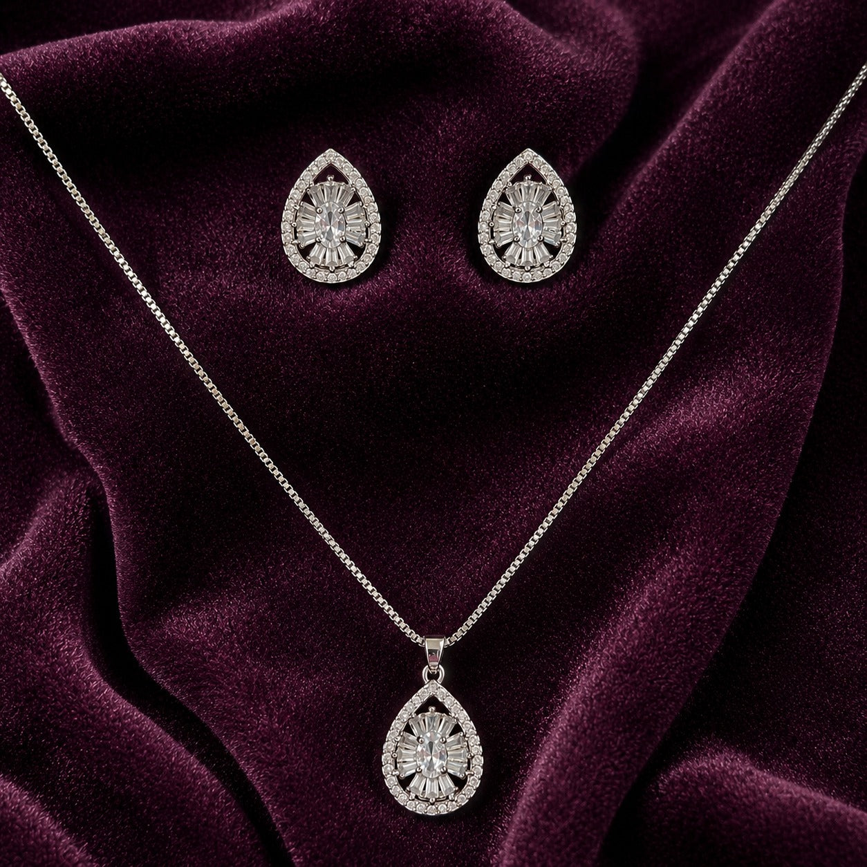 Dropline S925 silver earrings and pendant set with teardrop frames, clear zircon centres and baguette-style detailing displayed on deep plum velvet