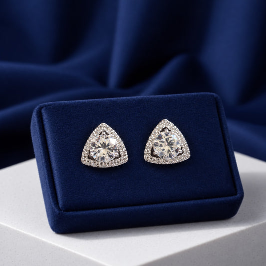 A pair of triangular stud earrings with clear gemstones in a silver-toned halo setting, displayed on a dark blue velvet jewelry box against a draped navy fabric background.