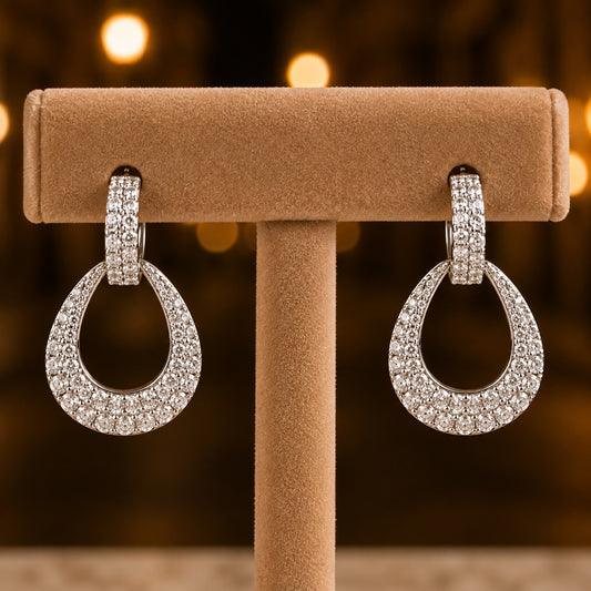 A pair of silver-toned teardrop-shaped drop earrings paved with clear stones, displayed on a tan jewelry stand against a warm, blurred bokeh background.