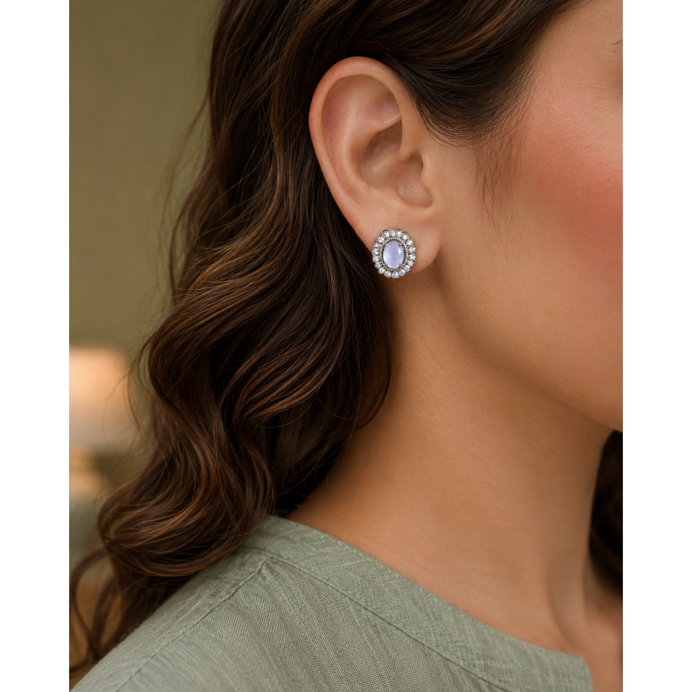 A close-up of a woman's ear wearing a round stud earring with a light blue oval center stone surrounded by a halo of small clear stones.