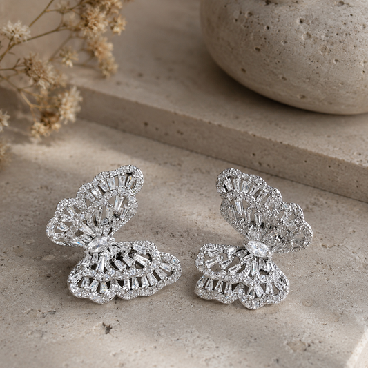 Luxury silver butterfly crystal earrings photographed on a premium travertine stone surface with soft studio lighting, highlighting the detailed openwork design and brilliant sparkle.