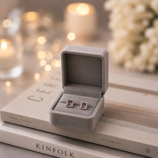 Square halo stud deep purple earrings displayed in a premium grey velvet jewellery box on stacked coffee table books with a softly blurred candlelit background.