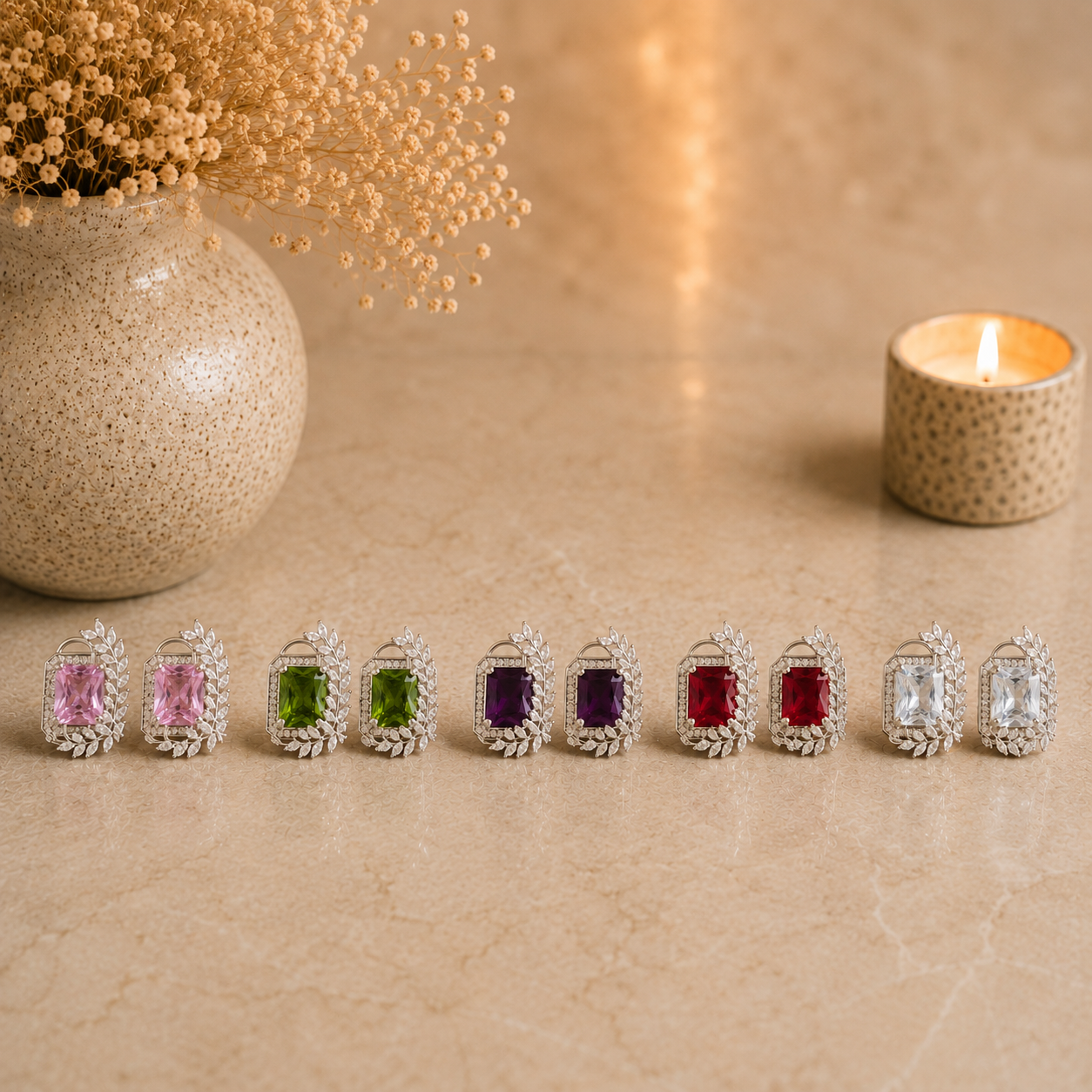 Five colour variants of silver leaf frame gemstone stud earrings displayed together on a warm beige marble surface with pink, olive green, deep purple, ruby red, and clear crystal stones. Luxury jewelry flat lay with a ceramic vase of dried flowers and a softly glowing candle in the background.