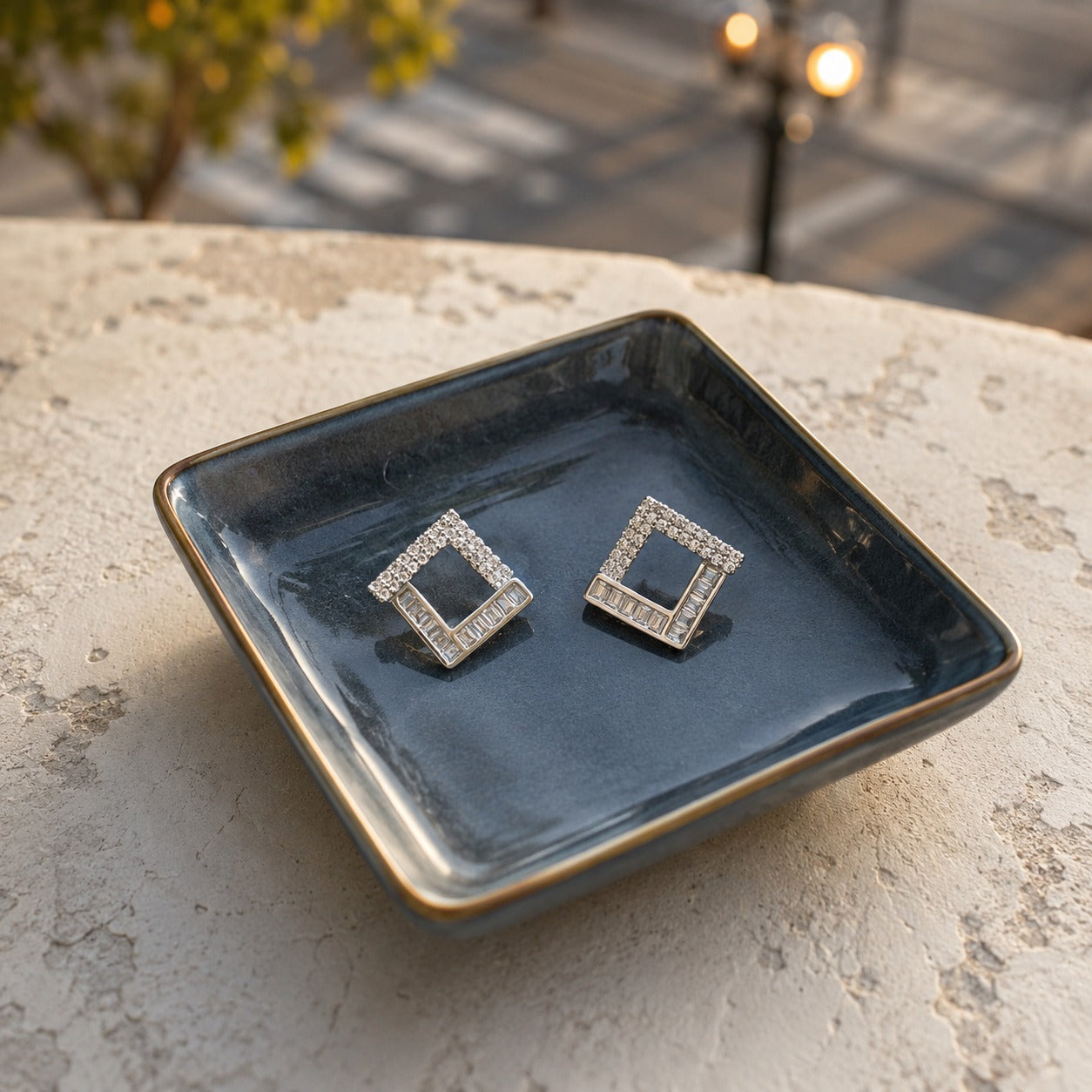 Silver geometric square crystal stud earrings with baguette-cut stones displayed on a premium ceramic tray in a luxury setting.
