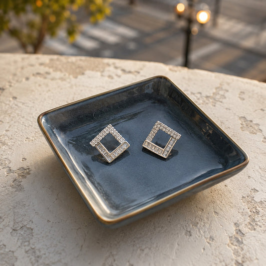Silver geometric square crystal stud earrings with baguette-cut stones displayed on a premium ceramic tray in a luxury setting.
