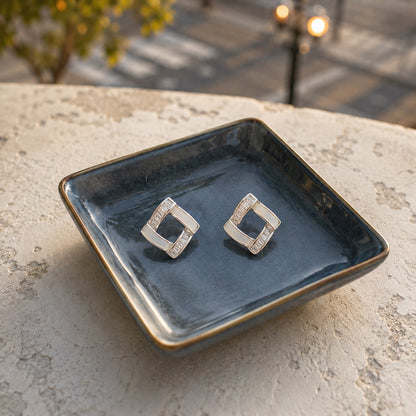 White geometric square crystal stud earrings with baguette-cut stones displayed on a premium ceramic tray in natural daylight